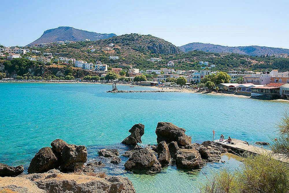 A sandy beach is bordered by clear turquoise waters, with gentle waves lapping against smooth rocks. In the background, a coastal town is visible, featuring white buildings nestled against a lush green hillside. Mountains rise in the distance under a clear blue sky.
