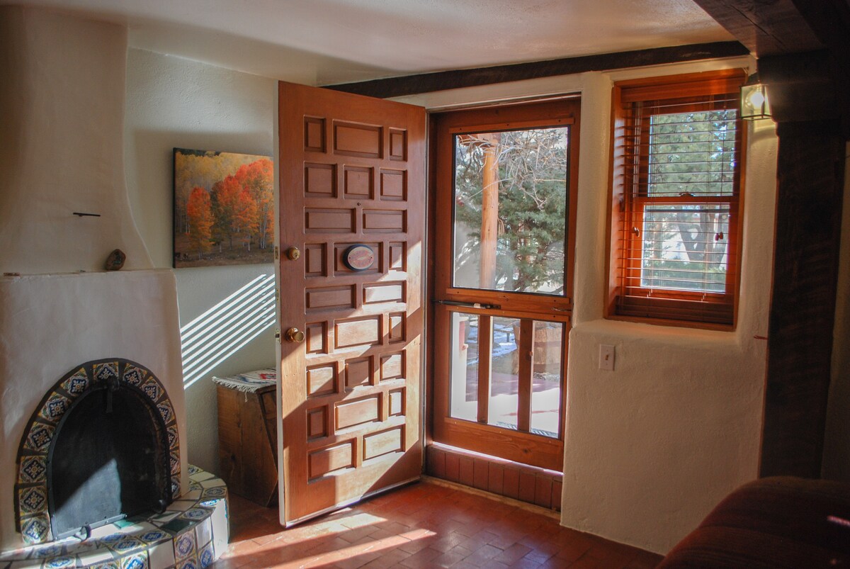 An inviting entryway features a wooden door with intricate paneling, welcoming light from a nearby window with wood blinds. A stone fireplace with decorative tiles stands beside a small table, and a vibrant piece of artwork adorns the wall.