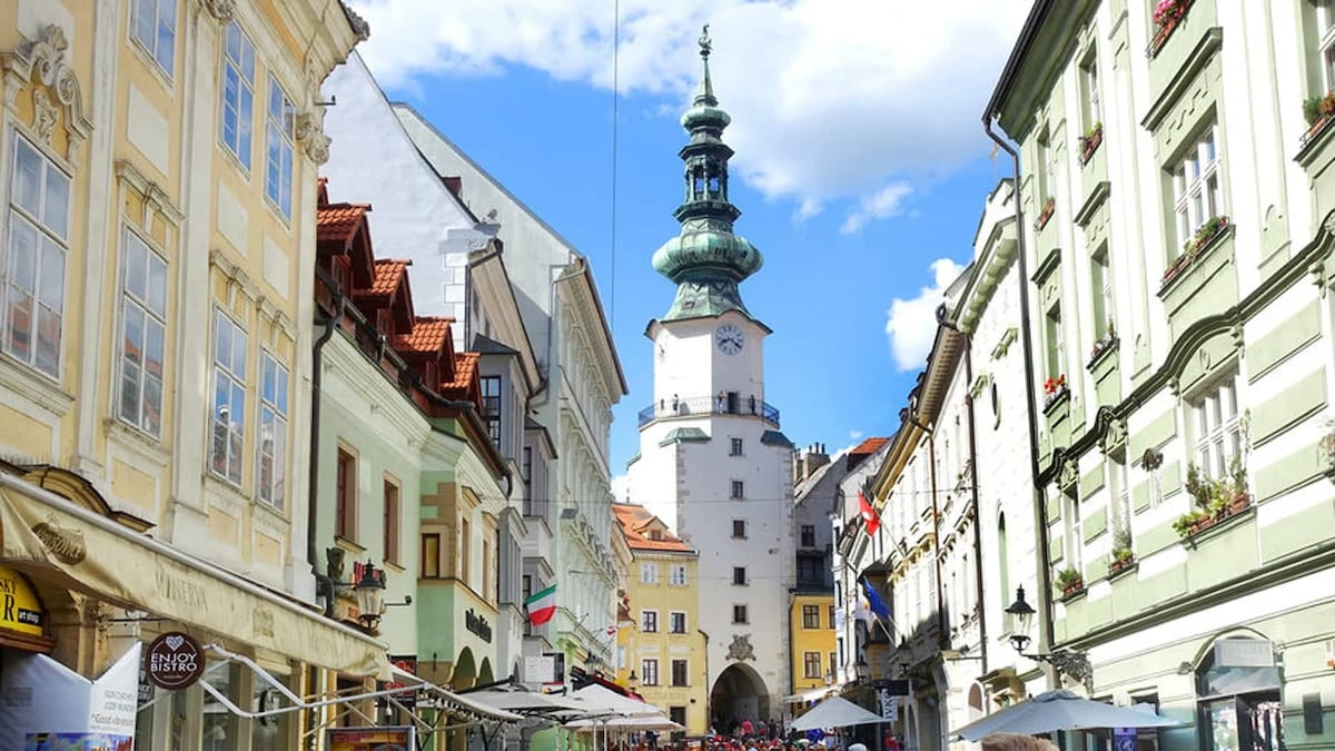A vibrant street scene in Old Town features charming buildings with varying architectural styles. A tall clock tower rises in the background, flanked by shops and outdoor cafés. Colorful flags are displayed, and the sky is bright with scattered clouds.