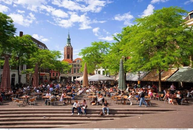 A vibrant square is filled with tables and chairs, occupied by individuals enjoying their time outdoors. Lush green trees provide shade, and a historic clock tower is visible in the background. The relaxed atmosphere is complemented by a clear blue sky dotted with clouds.