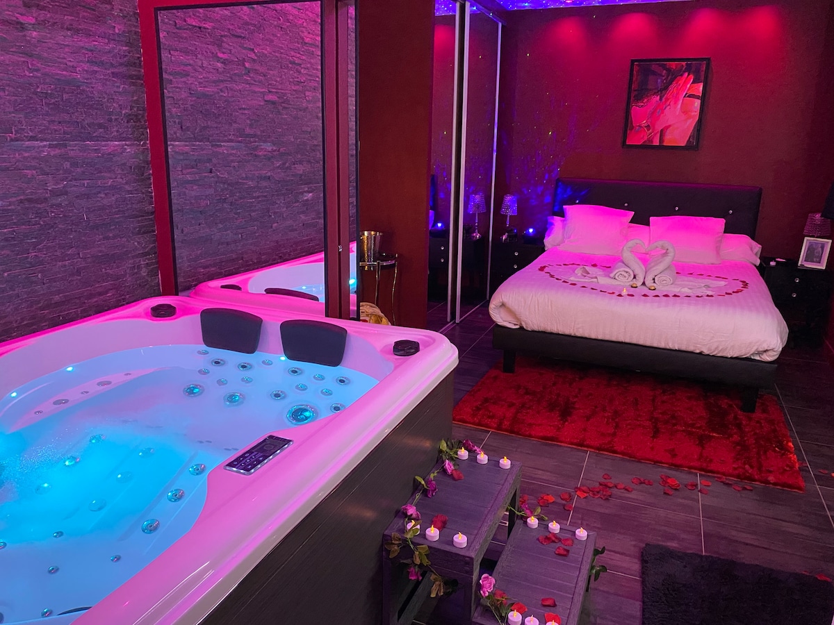 A serene space features a private jacuzzi illuminated with vivid blue and pink lights beside a elegantly decorated bed. Soft petals lie on the floor, creating a romantic ambiance, while a mirror reflects the mood lighting. The room offers a combination of relaxation and intimacy.