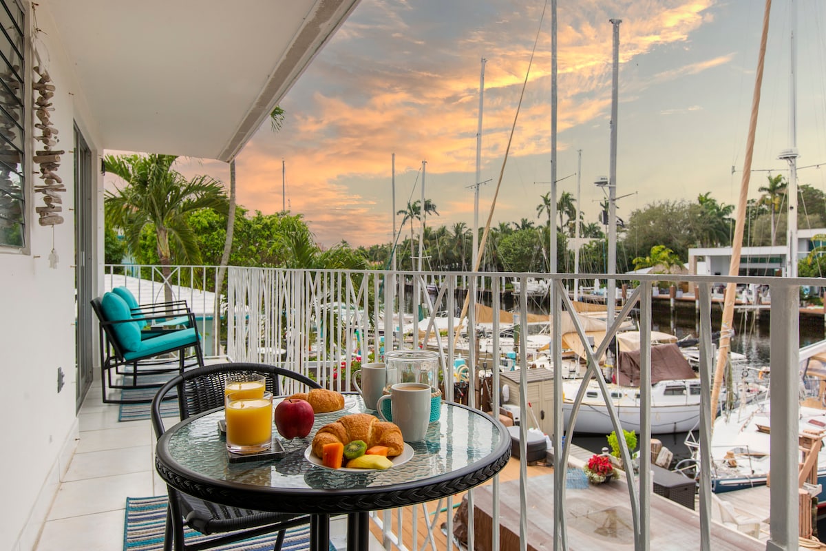 A private balcony is seen featuring a round table set with a breakfast spread, including a glass of juice, a cup, an apple, and croissants. Sailboats are docked in the waterway below, framed by trees and a vibrant sunset sky.