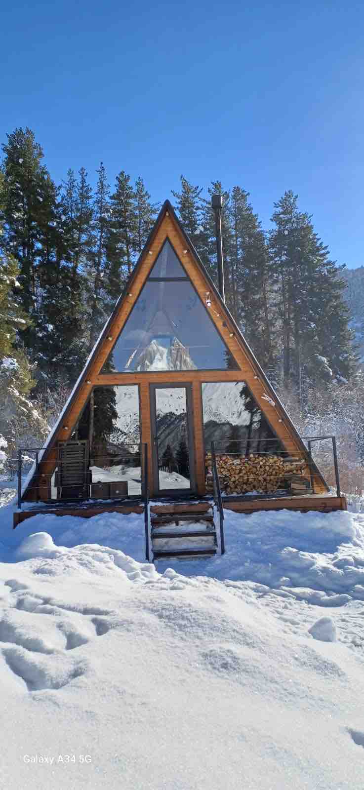 A modern A-frame cottage features a large glass front, allowing natural light to fill the interior. The structure is surrounded by snow-covered terrain and tall trees. A staircase leads up to the entry, with neatly stacked firewood visible beneath the porch.