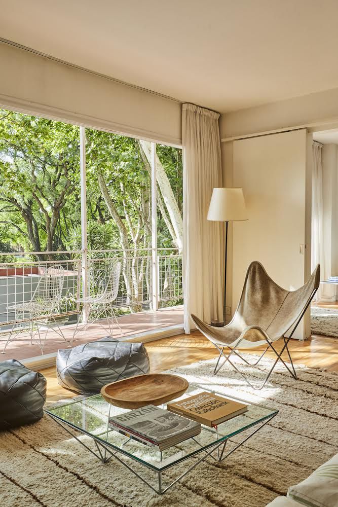 The living area features large glass doors that open to a balcony, allowing natural light to flood the room. A stylish butterfly chair and bean bag seating options are included, along with a glass coffee table displaying books and decorative items. The lush greenery outside enhances the ambiance.