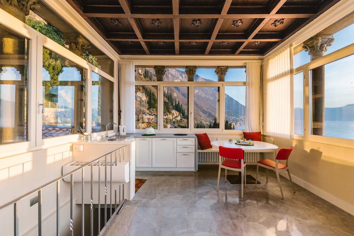 A bright dining area features a round table accompanied by four chairs, with views of the surrounding mountains and lake visible through expansive windows. The kitchen is equipped with modern appliances, and light floods the space through the large windows and skylights.