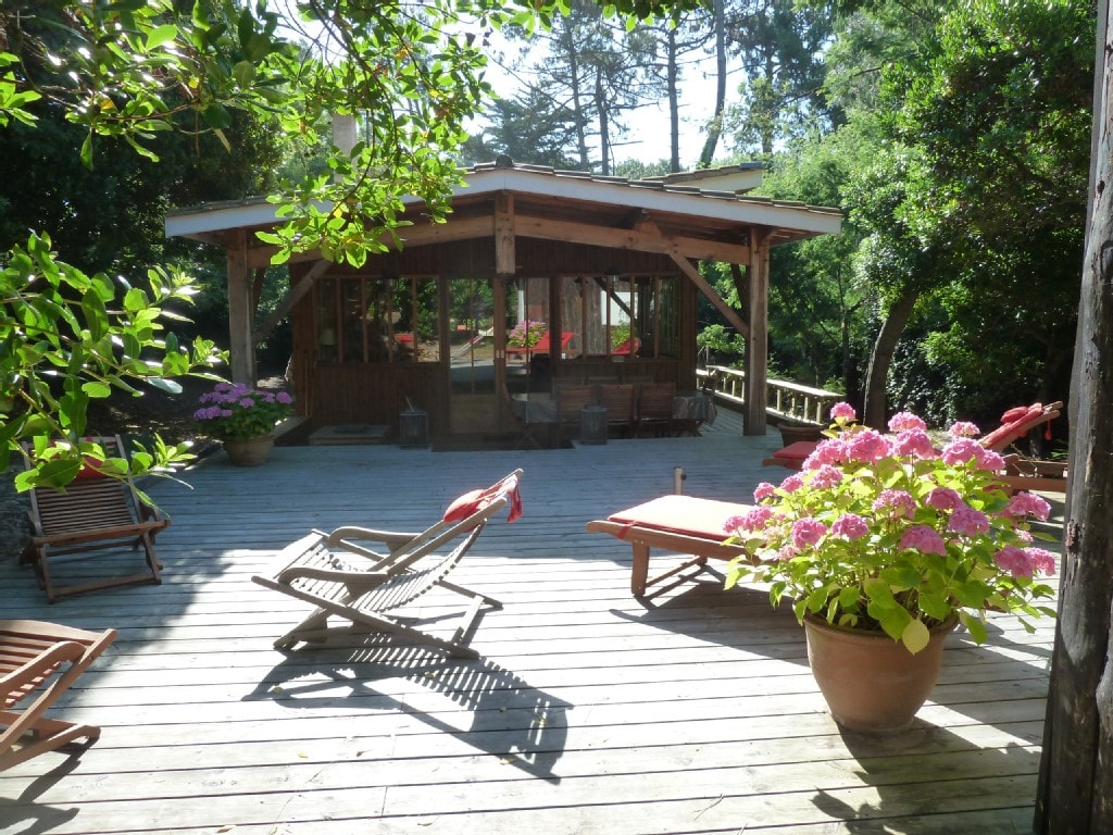 A spacious wooden deck is surrounded by vibrant foliage and blooming hydrangeas. Lounge chairs are arranged for relaxation, with a wooden structure visible in the background, featuring large glass windows that allow natural light to fill the interior.