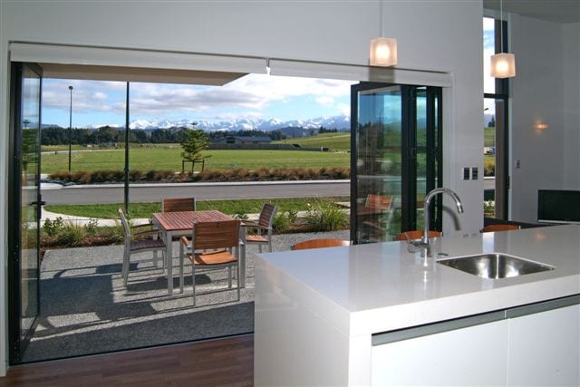 A modern kitchen with a sleek white countertop overlooks an outdoor dining area. Large glass doors open to a scenic view of rolling fields and mountains, inviting natural light into the space. Outdoor furniture is visible, set against a backdrop of greenery.