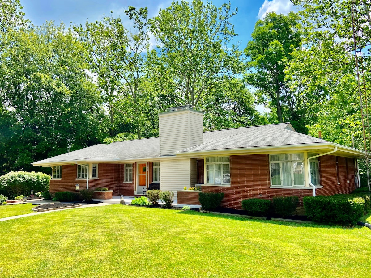 A charming Mid-Century house features a red brick exterior and a neatly landscaped front yard. Large windows provide natural light, and a central chimney accents the roof. Lush greenery surrounds the structure, creating a peaceful and inviting setting.