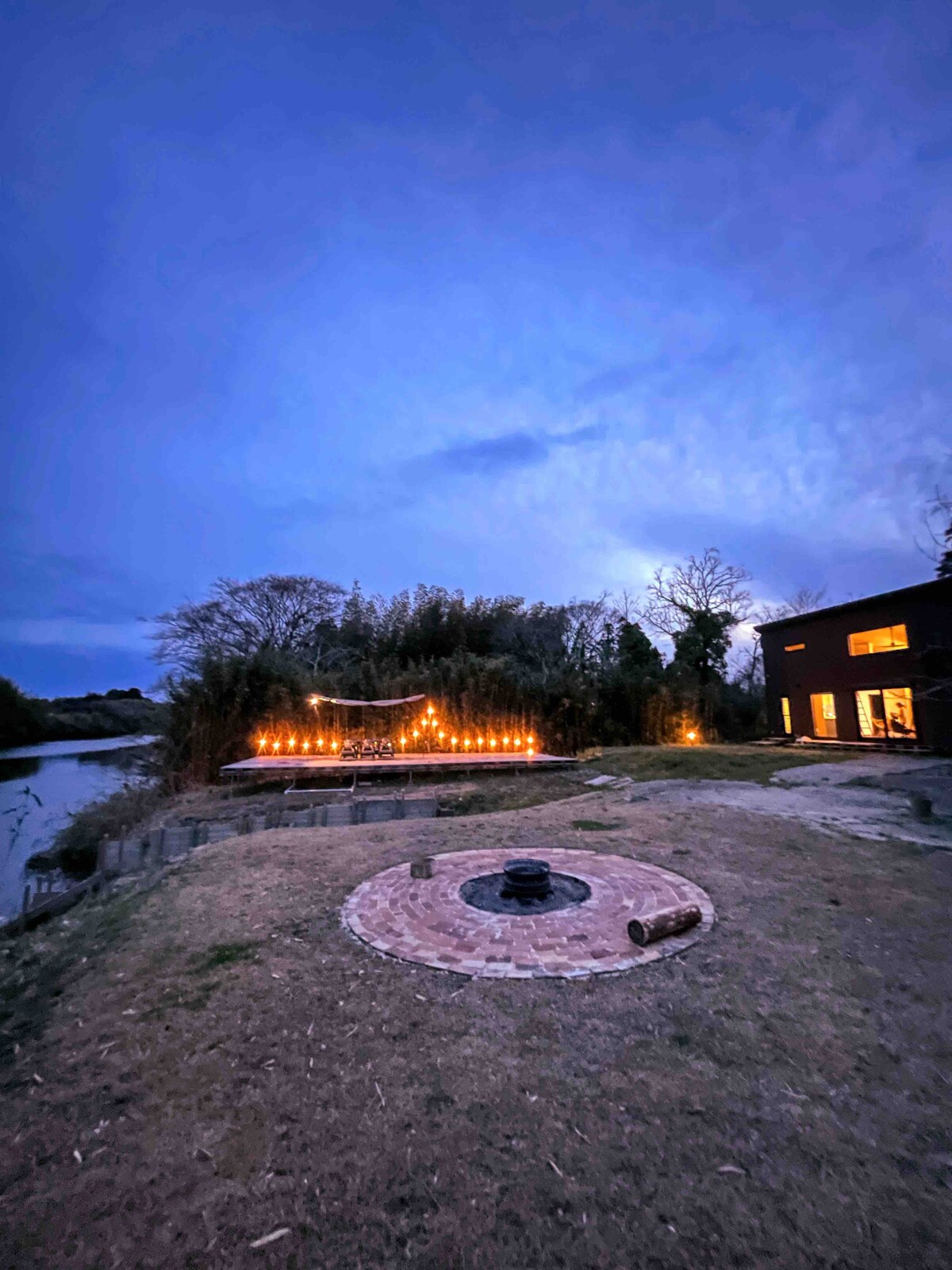 An outdoor fire pit surrounded by a circular brick patio is positioned on a grassy area, with a river softly reflecting the twilight sky nearby. Strings of lights are draped above a deck, creating a cozy ambiance for evening gatherings.