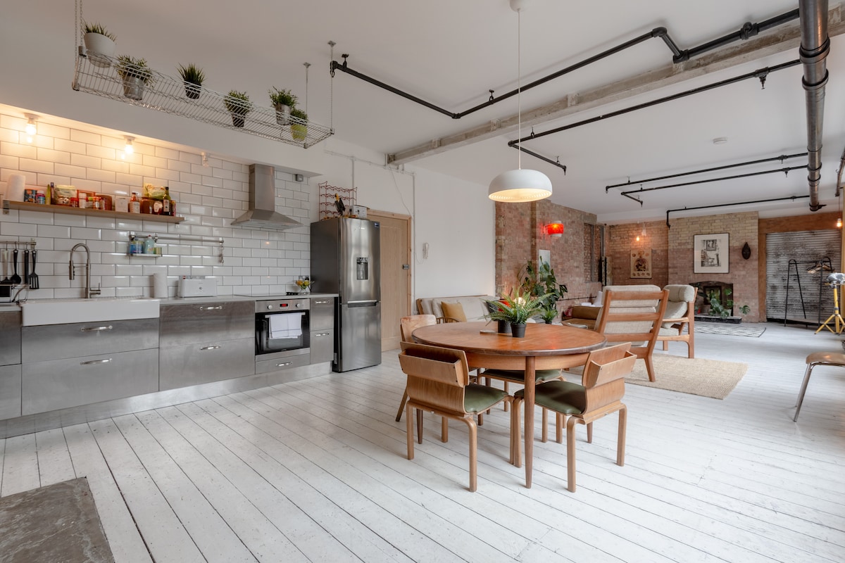 The open-plan kitchen and dining area features a modern kitchen with stainless steel appliances and white subway tile backsplashes. A wooden dining table is surrounded by light wood chairs, with plants adorning shelves above. Exposed brick and a large window provide an airy feel.