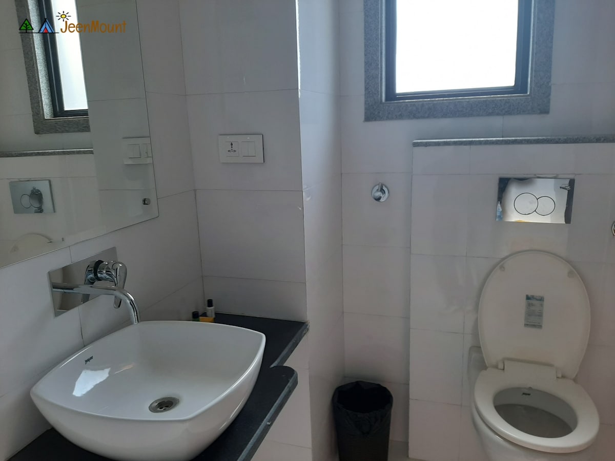 A modern bathroom is equipped with a sleek sink and large mirror, accentuated by natural light from the window. A toilet is located beside it, and a waste bin sits on the floor. The space is designed for functionality and comfort.