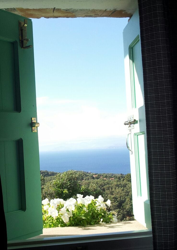 Open shutters frame a scenic view of the Ionian Sea and the surrounding hills. A window box filled with white flowers adds a touch of nature, while the sky showcases a mix of clouds and sunshine. The vibrant color of the shutters enhances the inviting atmosphere.