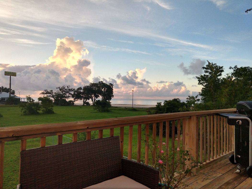 A wraparound deck features outdoor seating, providing a view of a tranquil landscape. Lush greenery and distant trees are visible, while soft clouds reflect warm hues in the sky above the calm waters of the Gulf.