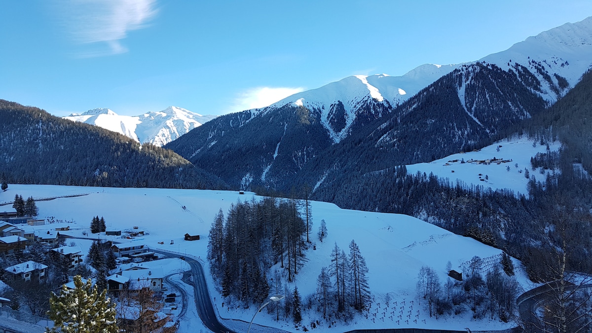 A panoramic view of snow-covered mountains and valleys is captured under a clear blue sky. Gentle slopes are adorned with trees, while quaint wooden structures are visible in the distance, contributing to the serene winter landscape.