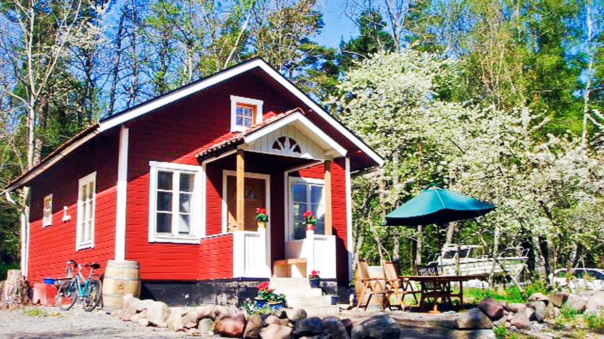 A charming red cabin with white trim is surrounded by blooming trees. A small outdoor seating area features a green umbrella and wooden furniture. A bicycle leans against the side of the cabin, and an inviting entrance with flower boxes completes the tranquil setting.