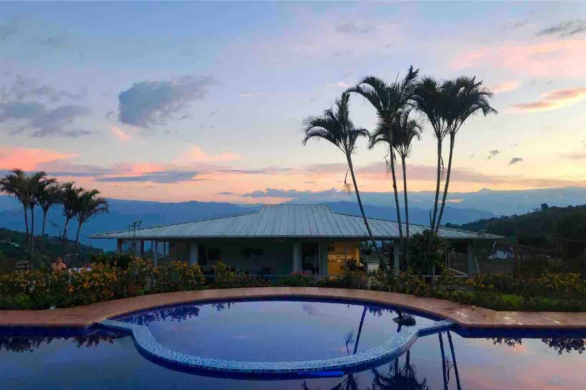 The image features a serene pool surrounded by lush greenery, with palm trees silhouetted against a colorful sunset sky. A spacious house is visible in the background, blending harmoniously with the natural landscape.