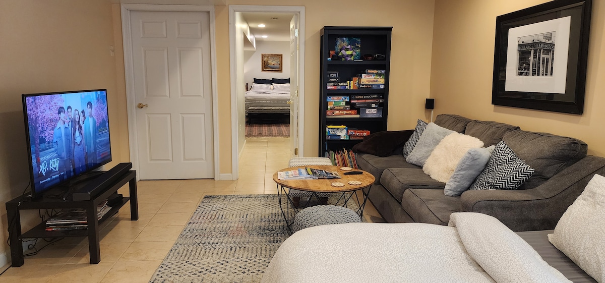 A comfortable living area features a gray couch adorned with decorative pillows, a round coffee table, and a large UHDTV displaying a show. The space is well-lit, with a doorway leading to a separate bedroom visible in the background.
