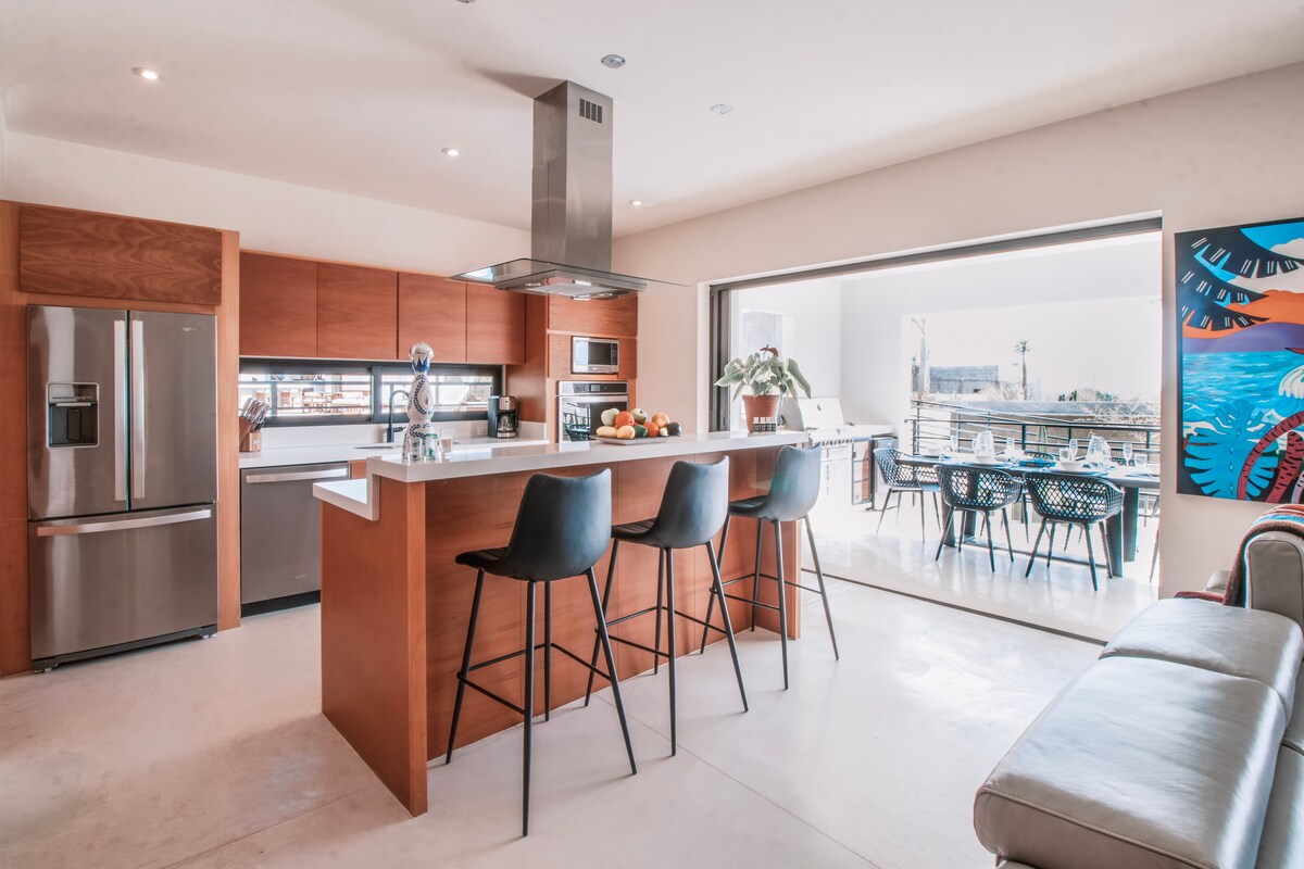 A modern kitchen features wooden cabinetry, stainless steel appliances, and a central island with seating. Natural light flows in through wide sliding doors, connecting the kitchen to an outdoor dining area. A vibrant piece of wall art adds a pop of color to the space.