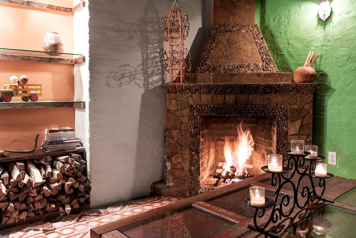 A warm stone fireplace is centrally positioned, with a fire gently glowing. Candles are placed on an ornate black table, creating a cozy ambiance. Stacked firewood is neatly arranged beside the fireplace, and decorative shelves adorn the wall, showcasing various items.