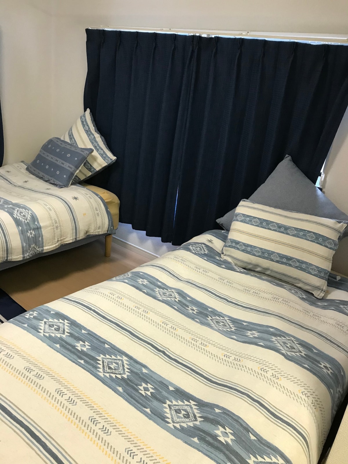 The room features two twin beds with patterned bedding in soft blue tones. Decorative pillows enhance comfort. Dark curtains cover the window, allowing for adjustable light control. A neutral-toned floor completes the cozy and inviting sleeping area.