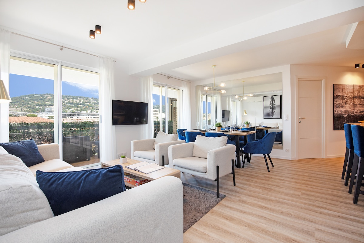 A spacious living and dining area features large windows offering views of the surrounding mountains. Comfortable seating includes a light-colored sofa and armchairs, complemented by a dining table set with blue chairs. Modern decorations and soft lighting create a welcoming environment.