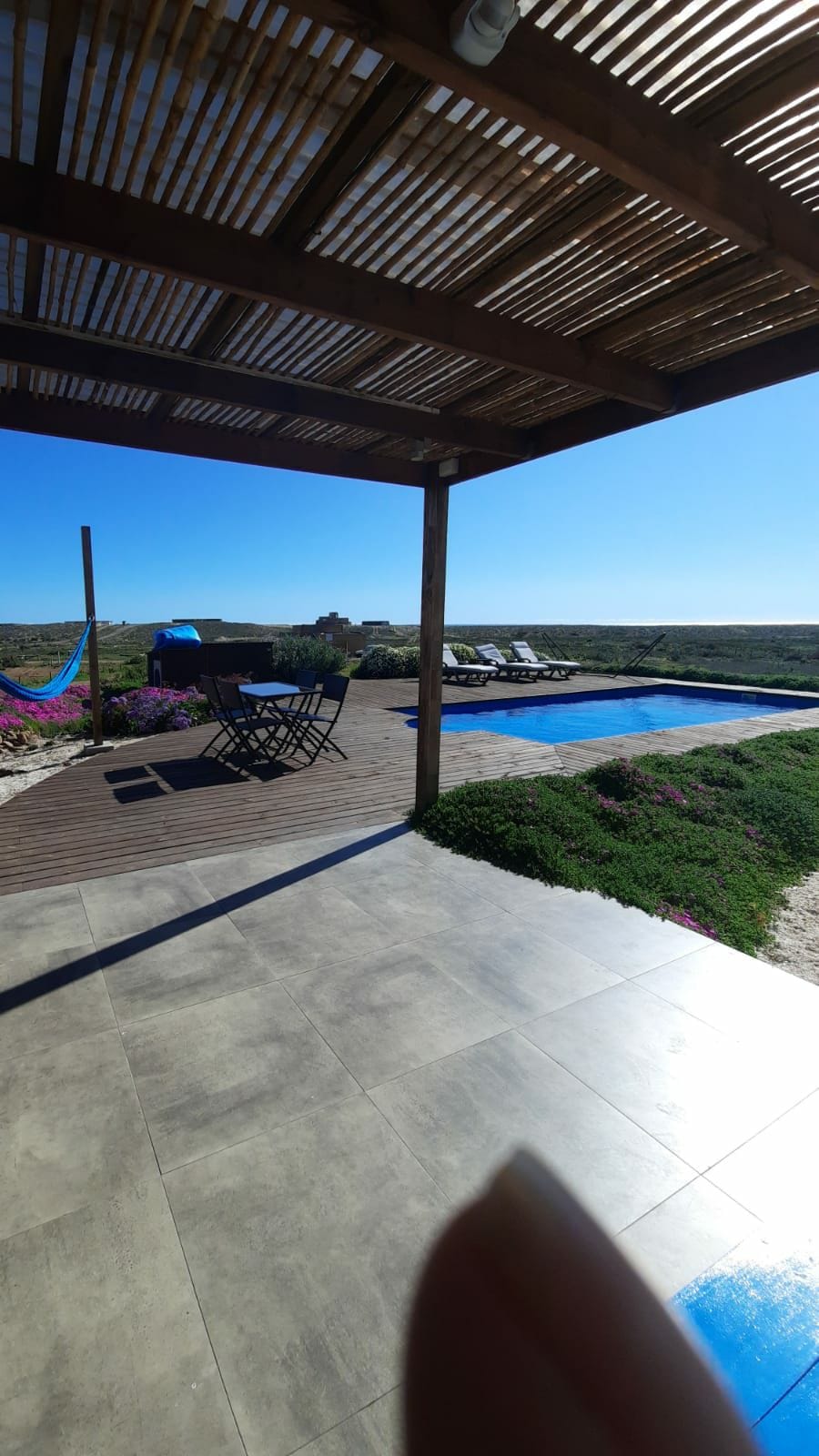 An outdoor terrace is featured overlooking a pool, surrounded by landscaped greenery. A dining set is positioned under a shaded area, accompanied by a hammock nearby. The bright blue sky enhances the serene atmosphere, and the pool reflects light in a calm manner.