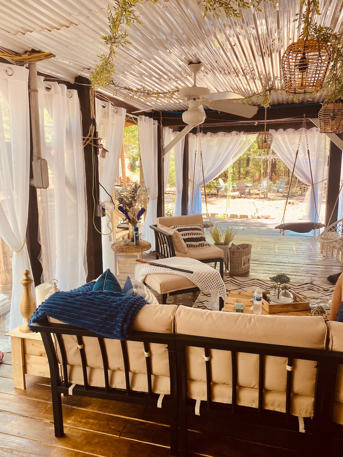 A cozy lounge area is adorned with light, flowing curtains and a ceiling decorated with natural elements. Comfortable seating is arranged with decorative pillows, inviting relaxation. A swing chair adds a playful touch, while the surrounding greenery is visible through the open layout.