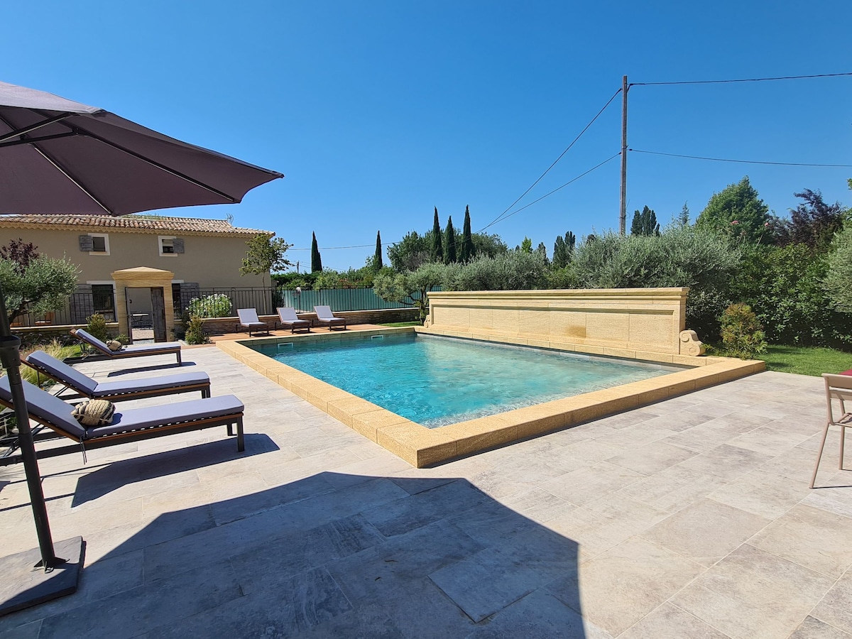 A spacious outdoor pool area features a sunlit swimming pool surrounded by sun loungers. Lush greenery and cypress trees create a serene backdrop, while a shaded seating area provides a comfortable spot for relaxation.