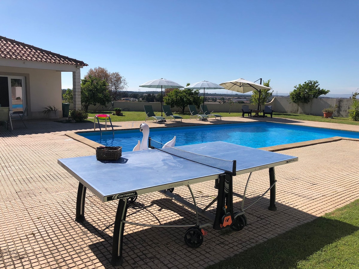 A ping pong table is positioned next to a sparkling blue pool, with lounge chairs and umbrellas surrounding the area. The scene is set against a clear blue sky, and lush greenery is visible in the background, enhancing the outdoor ambiance.