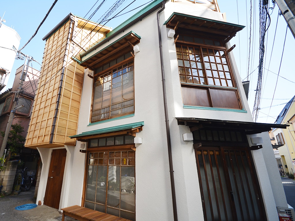 The exterior of a Japanese-style three-storied row house is showcased, featuring a combination of wooden and white plaster elements. Large windows with traditional grid patterns are present, and a small wooden bench is positioned at the entrance, enhancing the inviting ambiance.