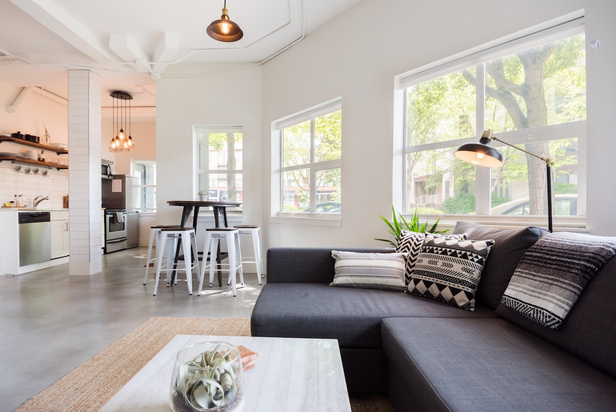 A modern living area features a spacious sectional sofa adorned with patterned cushions. Natural light streams through multiple windows, illuminating a small dining table surrounded by white stools. A minimalist coffee table with a decorative centerpiece sits nearby, adding to the contemporary aesthetic.