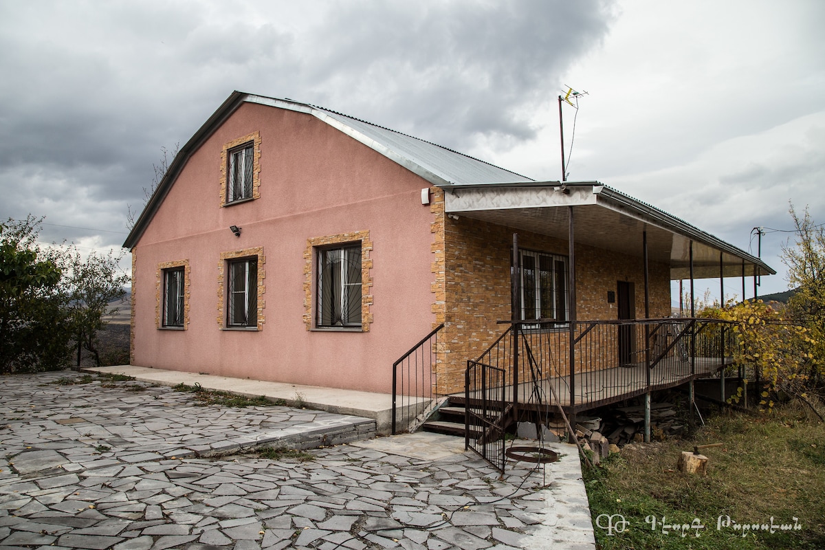The exterior of the Dzoragyugh Vacation Home features a two-tone facade with pink and brick elements. A covered porch runs along the front, providing access to the main entrance. Surrounding landscapes include views of greenery and a rocky pathway leading up to the home.