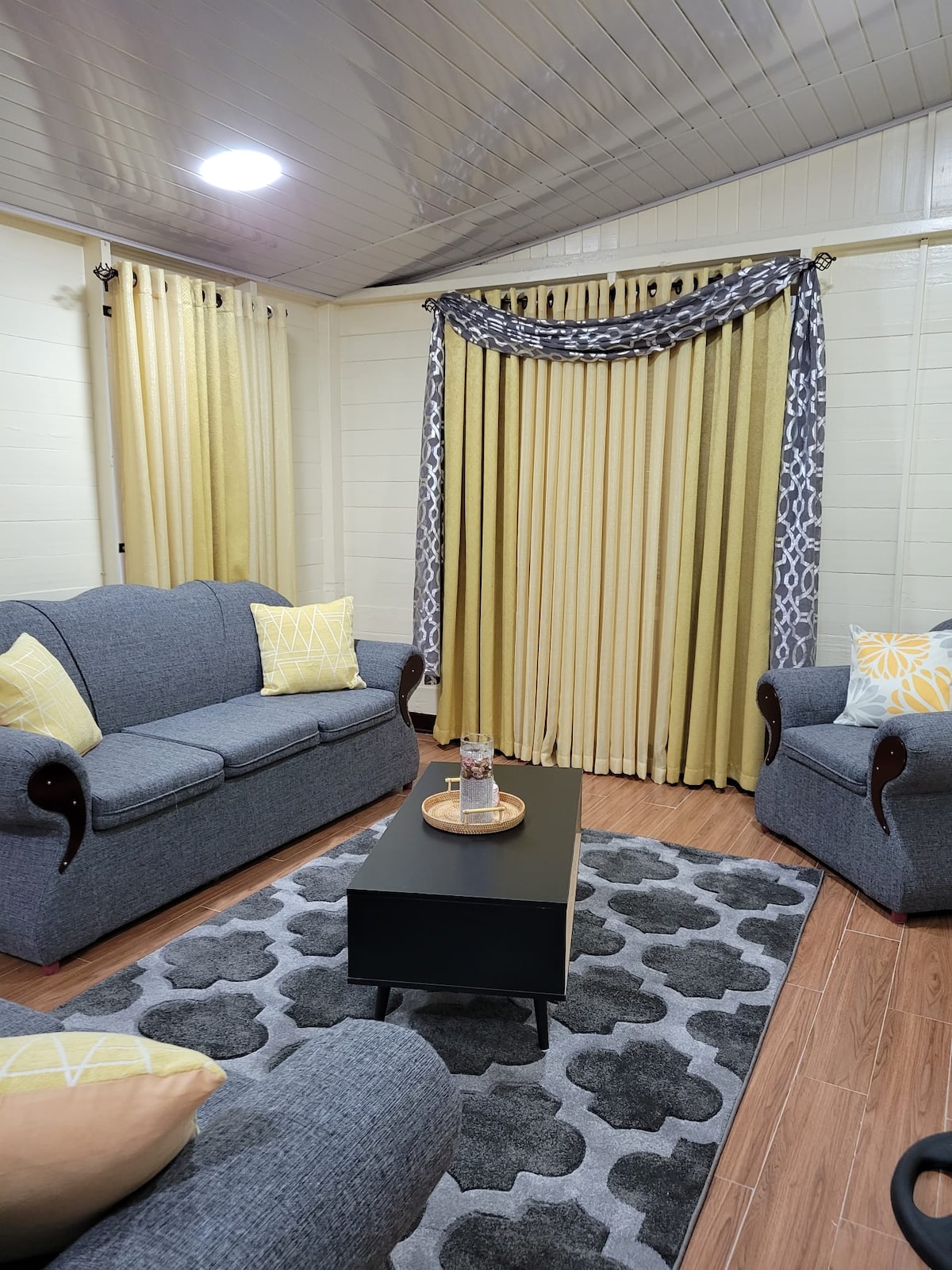 The living room features a gray sofa and armchair, adorned with patterned yellow throw pillows. A rectangular coffee table with a tray sits at the center, surrounded by a plush area rug. Natural light filters through patterned curtains, enhancing the inviting ambiance.
