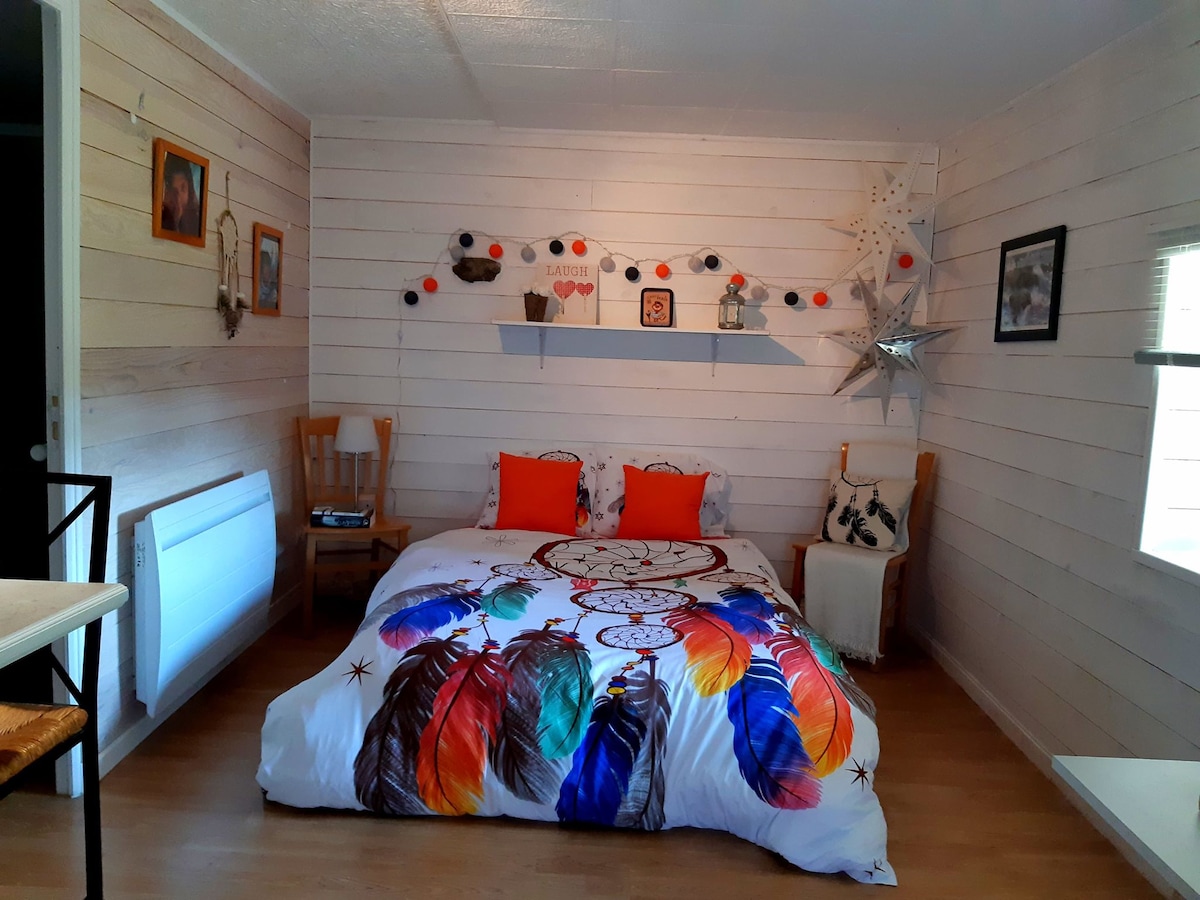 A comfortable sleeping area features a colorful bedspread with feather designs, complemented by bright orange pillows. The walls are lined with light wood paneling, and decorative elements, such as photographs and wall art, are thoughtfully arranged above the bed.