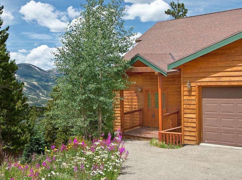 A well-maintained entrance of a luxury mountain home is framed by vibrant wildflowers and trees. The exterior features wood siding and a garage. Breathtaking mountain views are visible in the background under a clear blue sky.