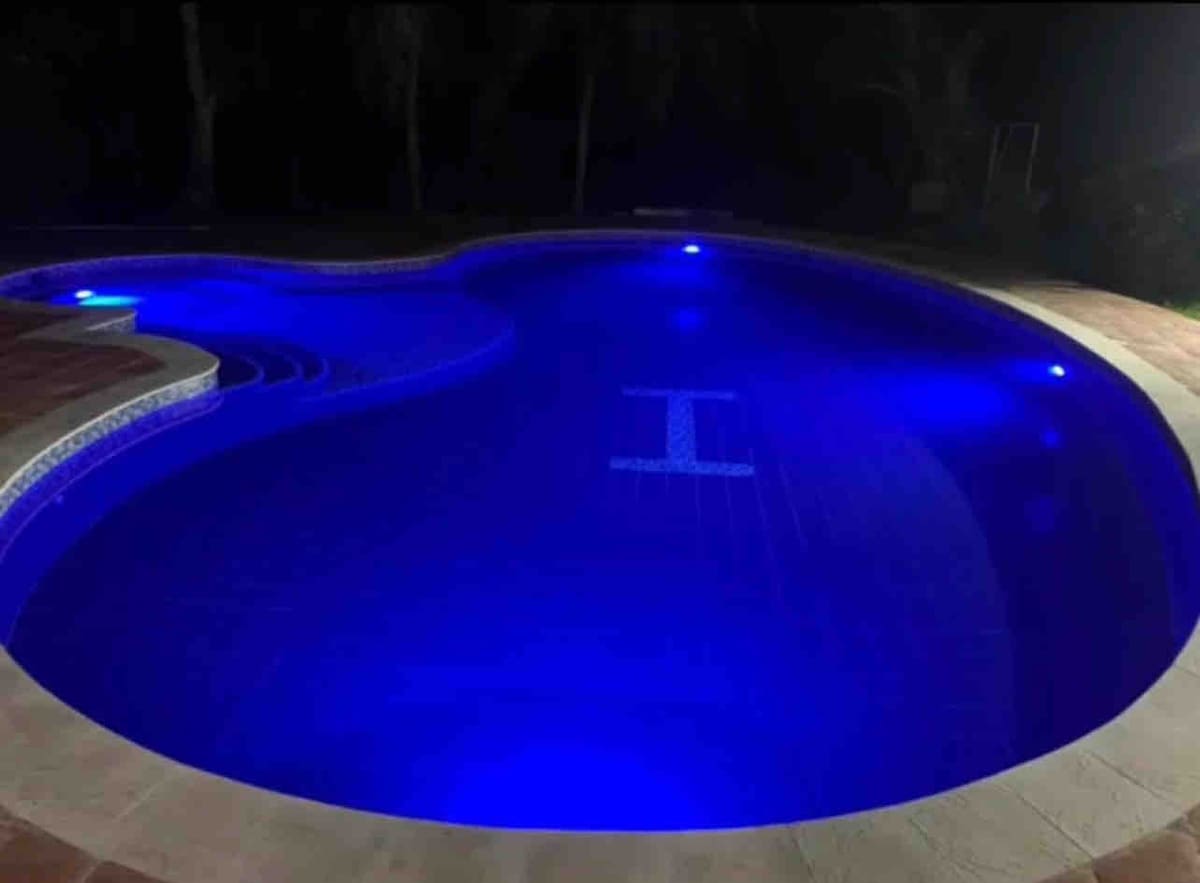 The pool features a unique shape, illuminated with soft blue lights, creating a serene environment. The water's surface reflects a tranquil ambiance, while the surrounding area includes tiled edges that enhance safety and ease of access.
