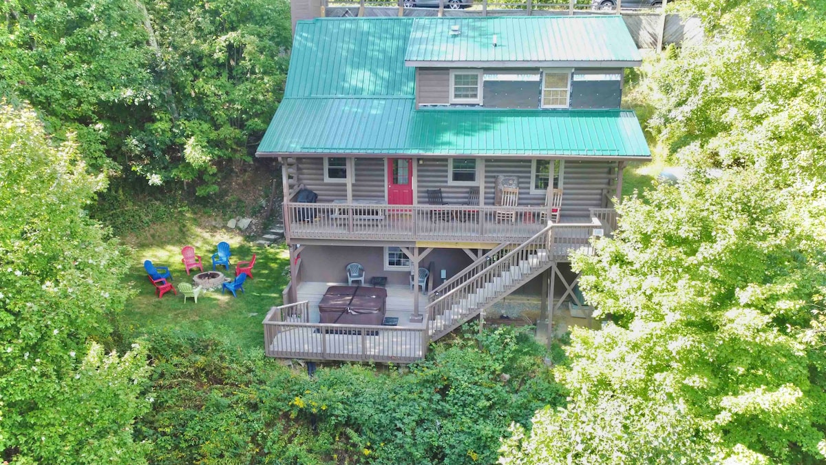 An aerial view of Bear Pause Cabin reveals a rustic log structure with a green metal roof. The spacious deck is equipped with a hot tub and surrounded by seating areas, framed by lush greenery, creating a serene outdoor setting.