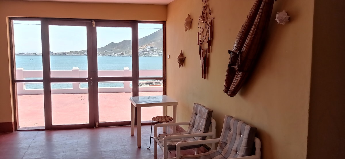 A bright and airy space features large glass doors that offer a view of the sea. Two light-toned chairs and a small round table are arranged on the red tiled floor, while decorative elements, including a wall hanging, complement the inviting atmosphere.