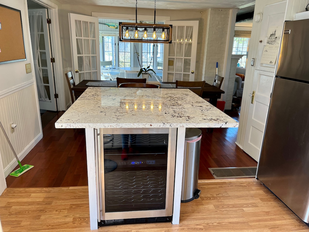 The kitchen features an elegant island with a granite countertop and bar seating. A wine cooler is integrated into the base, while overhead lighting provides a bright ambiance. The dining area is visible in the background, complemented by large glass doors leading to a sunlit space.