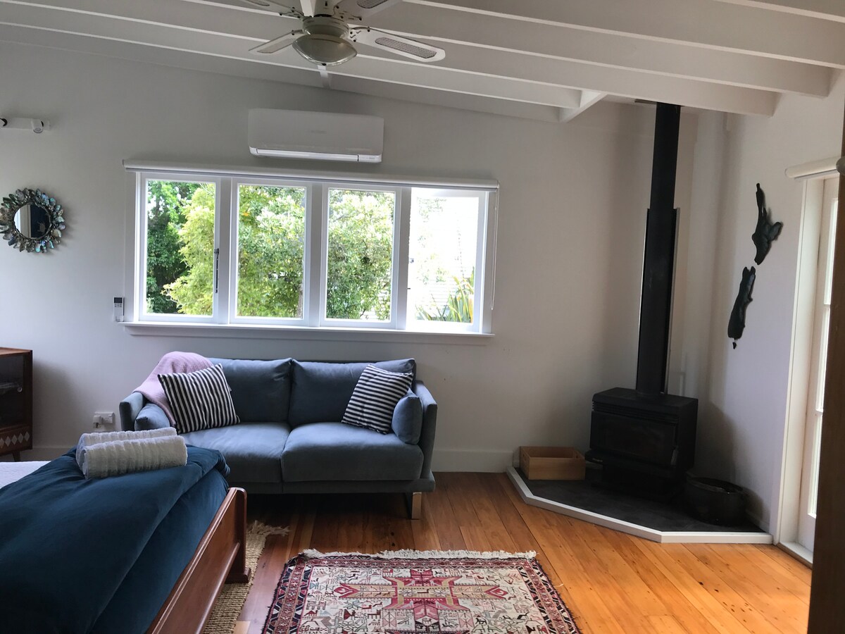 A cozy living area features a blue sofa adorned with striped cushions, positioned near large windows allowing natural light. A rustic rug covers the wooden floor, complemented by a wood-burning stove placed against the wall. A ceiling fan and wall art add character to the space.