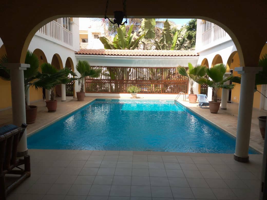 A clear view of a serene pool surrounded by lush greenery and palm trees. The spacious outdoor area features stone tile flooring and is framed by arched columns. Chaise lounge chairs are positioned nearby, offering relaxation options in a tranquil setting.
