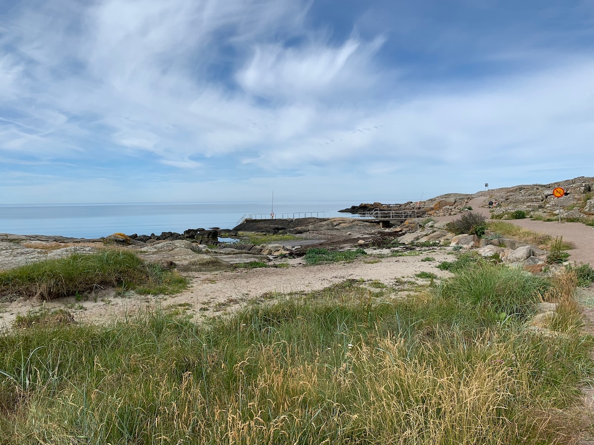 A coastal scene captures a rocky shoreline with gentle waves lapping against the rocks. A wooden pier extends into the water, while scattered grassy areas and paths are visible. The sky features soft clouds, enhancing the serene atmosphere of the landscape.