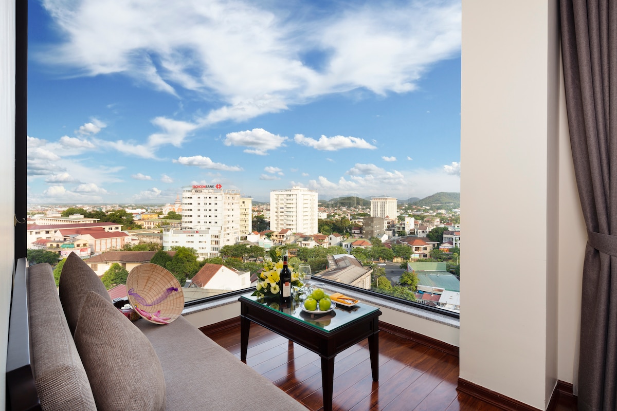 A spacious seating area is highlighted by a low table set with fruit and wine, offering panoramic views of the cityscape through large glass windows. Soft curtains frame the scene, while natural light brightens the polished wooden floor.
