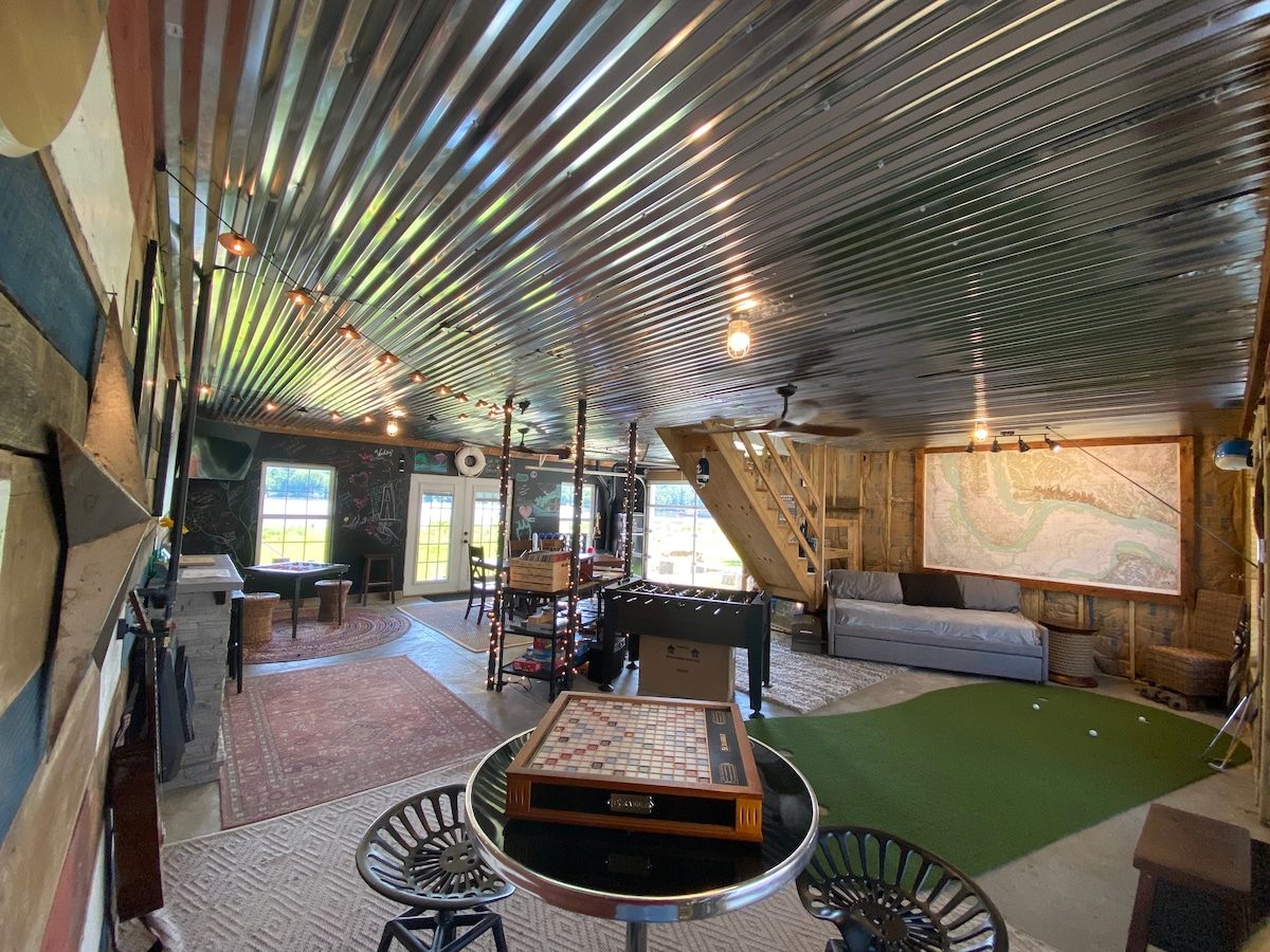 An open recreational space is depicted, showcasing a mix of games and seating. A putting green occupies the floor, accompanied by a foosball table. Exposed metal ceiling adds character, while large windows and a staircase provide natural light and access to upper areas.