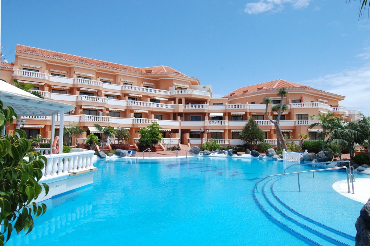 The image showcases a well-maintained pool area surrounded by a multi-level building. Sun loungers and palm trees line the inviting swimming pool, which features a gentle gradient. Clear blue skies are visible above, enhancing the serene vacation atmosphere.