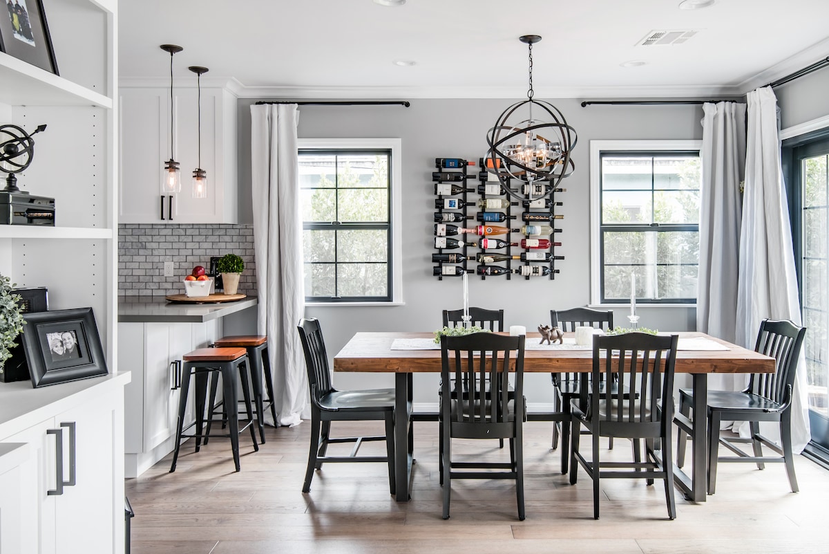 A modern dining area features a large wooden table surrounded by six black chairs. Two windows with light curtains offer natural light, while a wine rack is mounted on the wall, displaying various bottles. A small bar area with stools is visible in the corner.