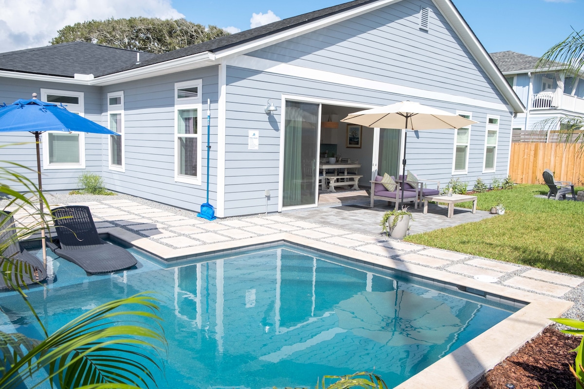 The backyard features a heated pool surrounded by a stone patio, with a couple of umbrellas providing shade. Lounge chairs are positioned near the water, and a dining area is visible through sliding glass doors leading into the home.