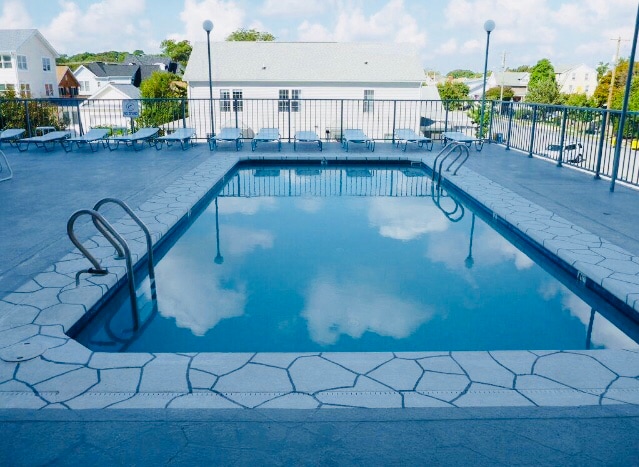 The outdoor pool area features a rectangular, inviting swimming pool surrounded by a textured stone deck. Several lounge chairs are positioned around the pool. The clear blue water reflects a partly cloudy sky, creating a serene atmosphere.