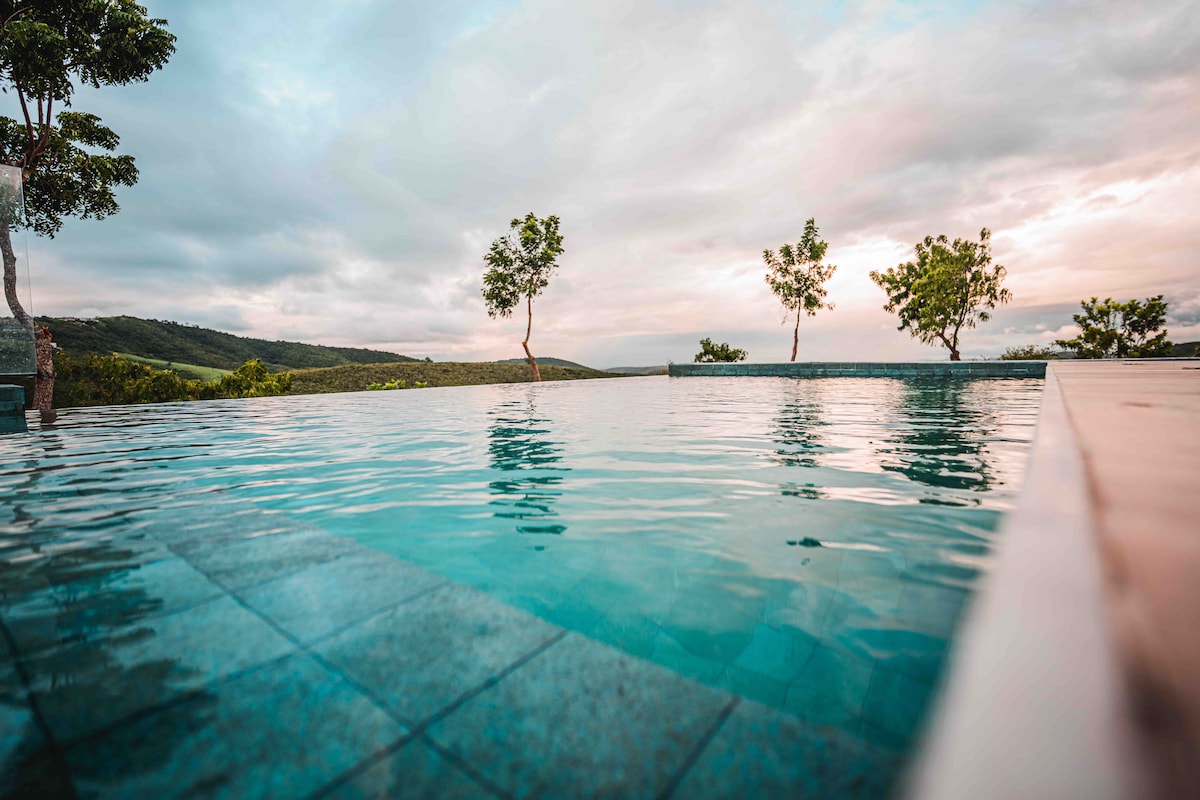 An infinity pool reflects the surrounding landscape, with gentle waves lapping at its edges. Green trees are positioned nearby, complementing the serene view of rolling hills and a colorful sky in the background.