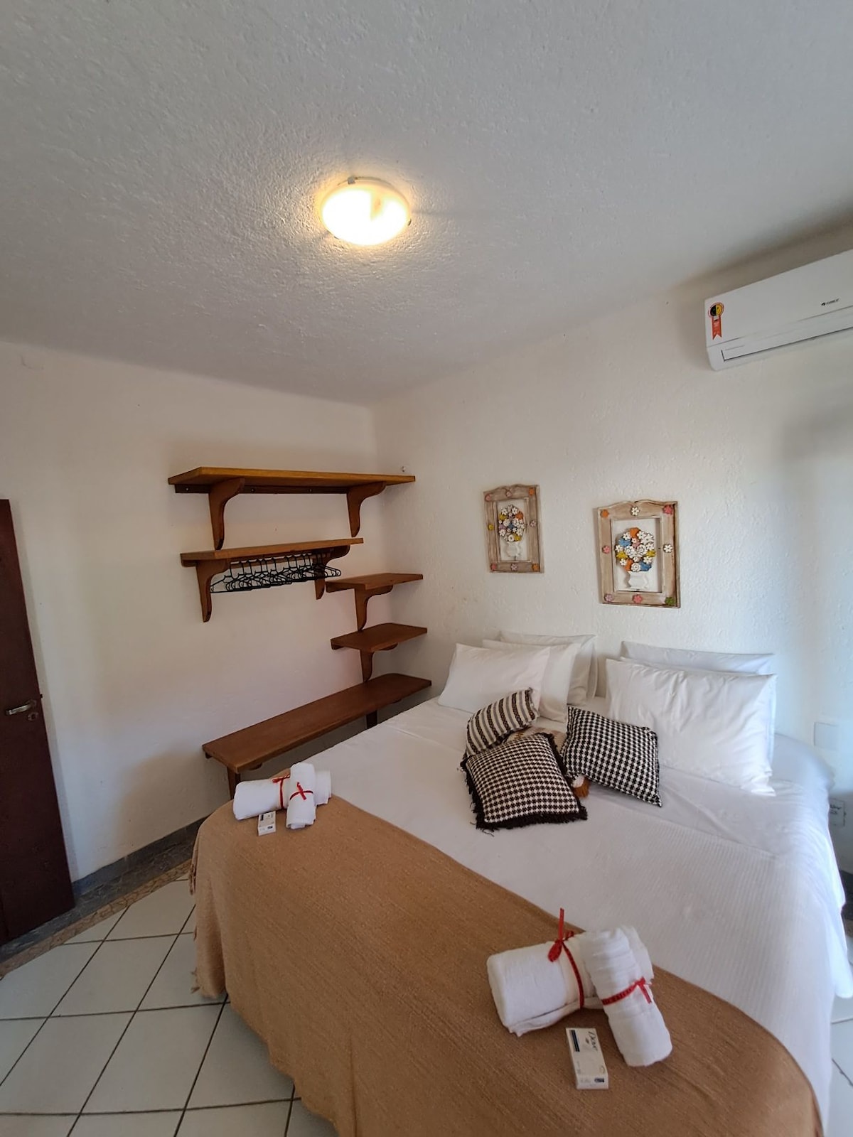 A comfortable suite features a spacious bed with neatly arranged pillows. A light-coloured blanket covers the bed, complemented by decorative cushions. Shelves with wooden finishes are mounted on the wall, while framed artwork adds character. Natural light fills the room through a nearby window.